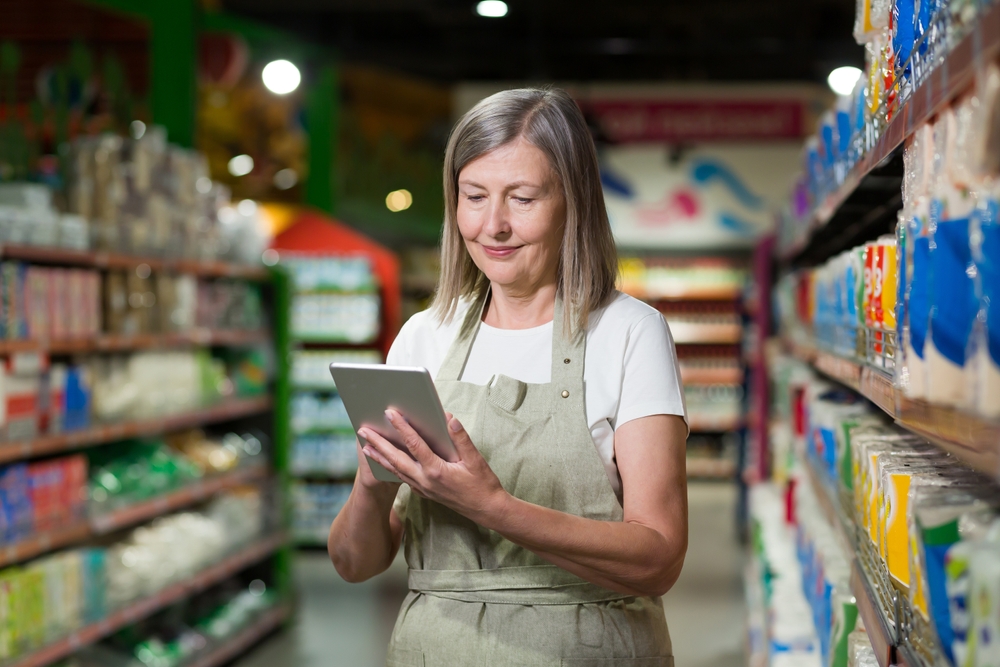 Mulher, branca, loira, com aproximadamente 50 anos, em um supermercado, trabalhando com um tablet nas mãos